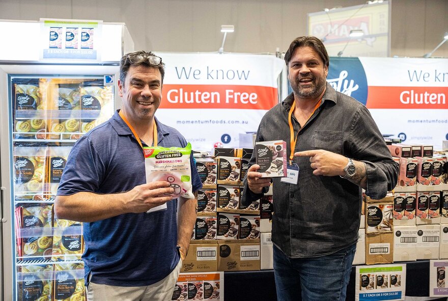 People interacting with an exhibition stallholder at the Gluten-Free expo, standing in front of a table that displays food products, with signage behind the stall.