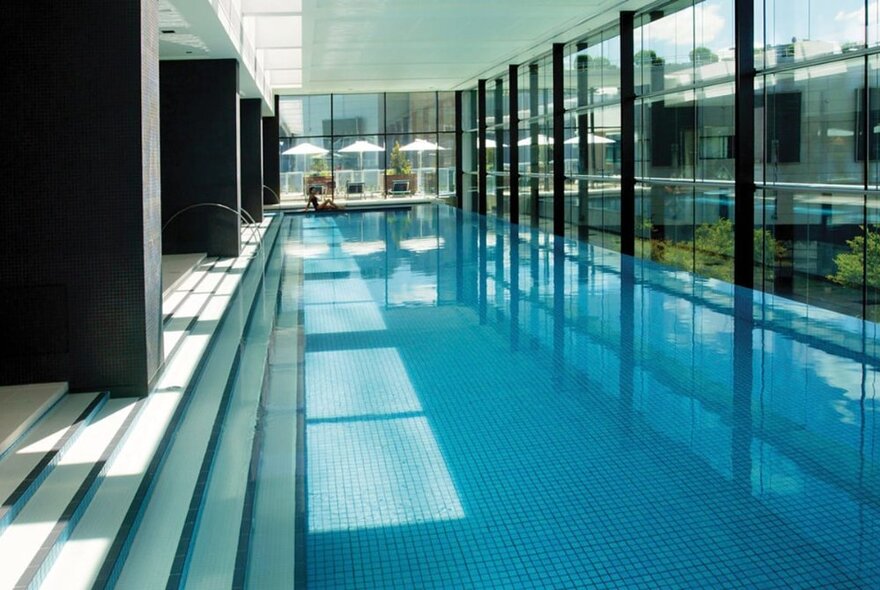 Long indoor hotel pool with blue tiles, steps leading into the pool along one side and windows looking outside on the other.