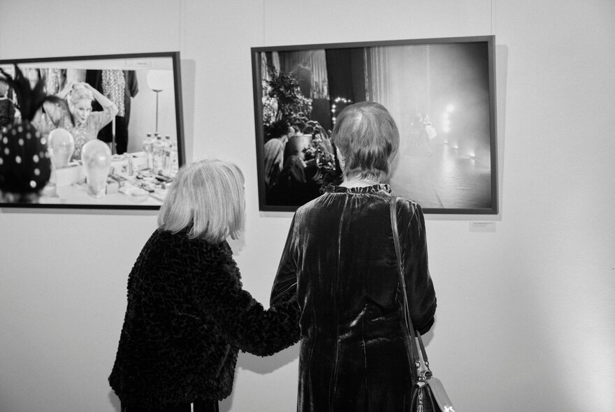 Black and white photograph of two women looking at a framed photograph hanging on a gallery wall.