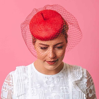A model wearing a frilly white shirt and a red pillbox hat and veil, against a pink background.