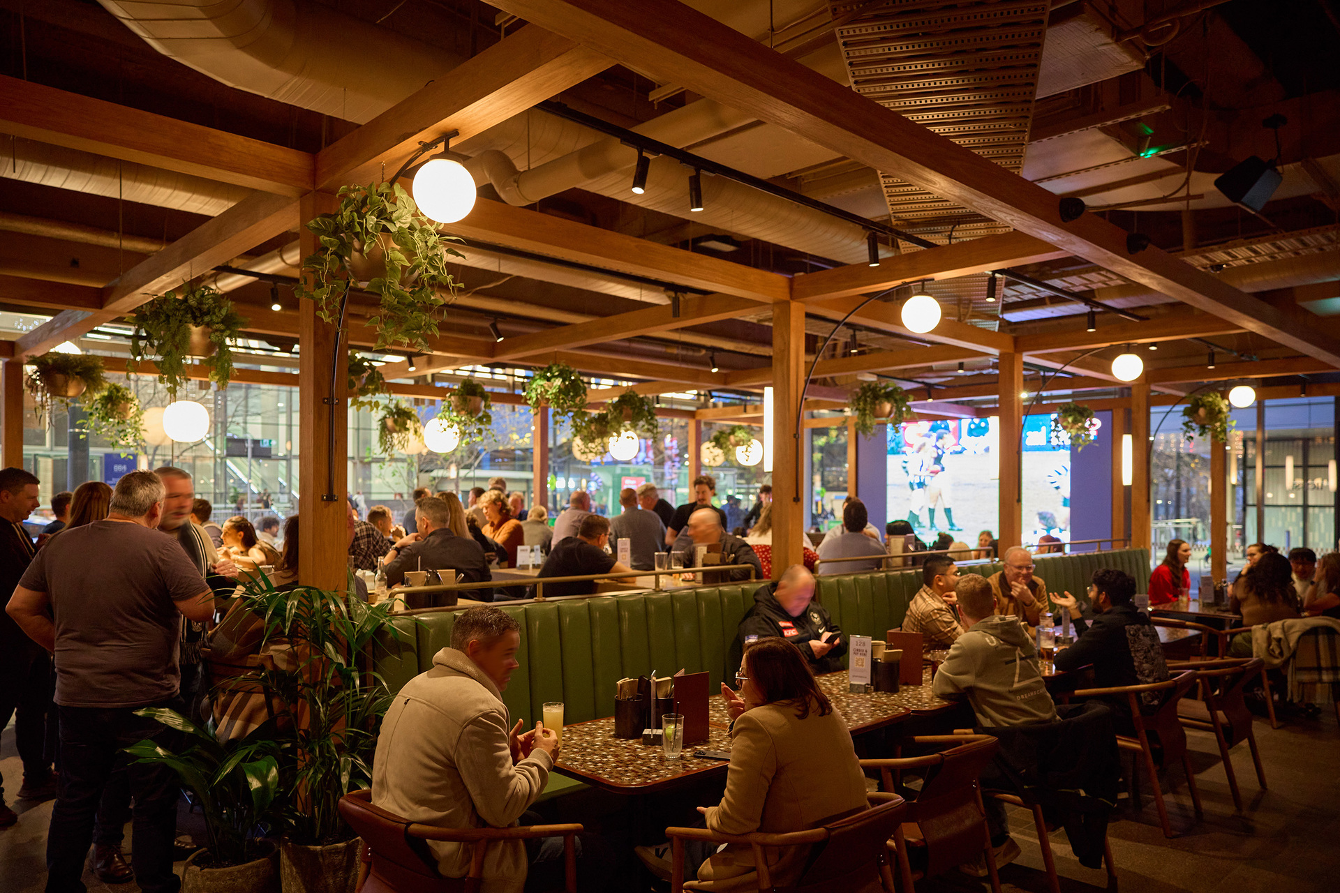 A crowd of people are inside a busy pub. Some are seated in booths while others are standing. There are hanging plants on the ceiling beams. 