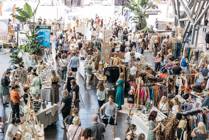 Aerial view of an interior market with stalls selling clothing and raft, and many people walking around and shopping in the space.