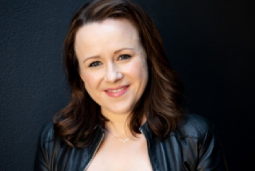 Portrait of a woman with brown hair, wearing a black leather jacket, in front of a black background.