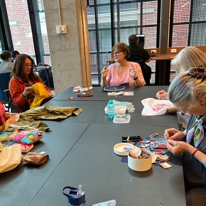 A group of people seated around a large table in a library space with sewing projects and materials. 