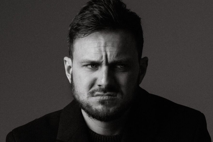 Man in his 30s, with short hair and a beard, his brow is furrowed and his eyes look to the side; black and white photo.