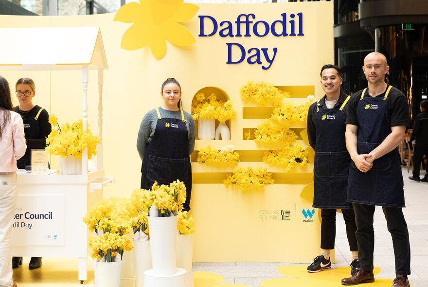 People wearing aprons standing at a display of masses of daffodils in white buckets at Collins Place retail square, a sign with the words Daffodil Day in the background.
