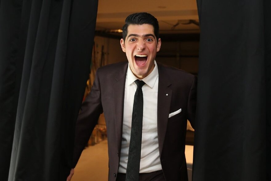 Singer William Le Brun wearing a suit and with his mouth open as if laughing, stepping through two stage curtains that he is pulling apart.