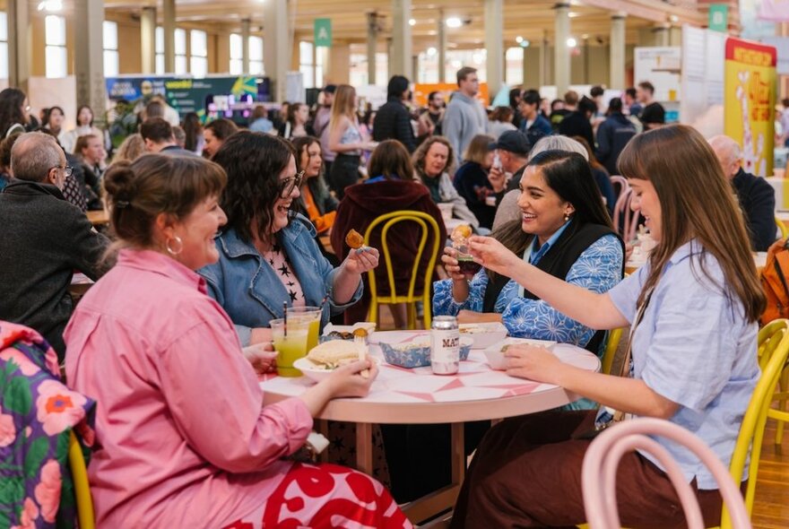 Table of four friends smiling and eating food in a large and crowded hall.