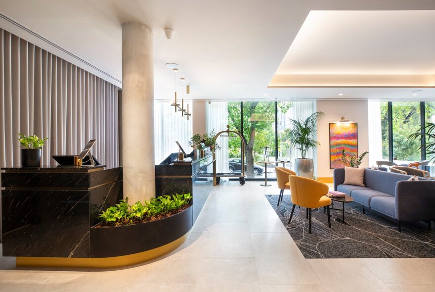 Hotel lobby featuring a curved front desk wrapped around a large concrete structural pillar, with sofas, chairs, lamps and green plants in the reception area.