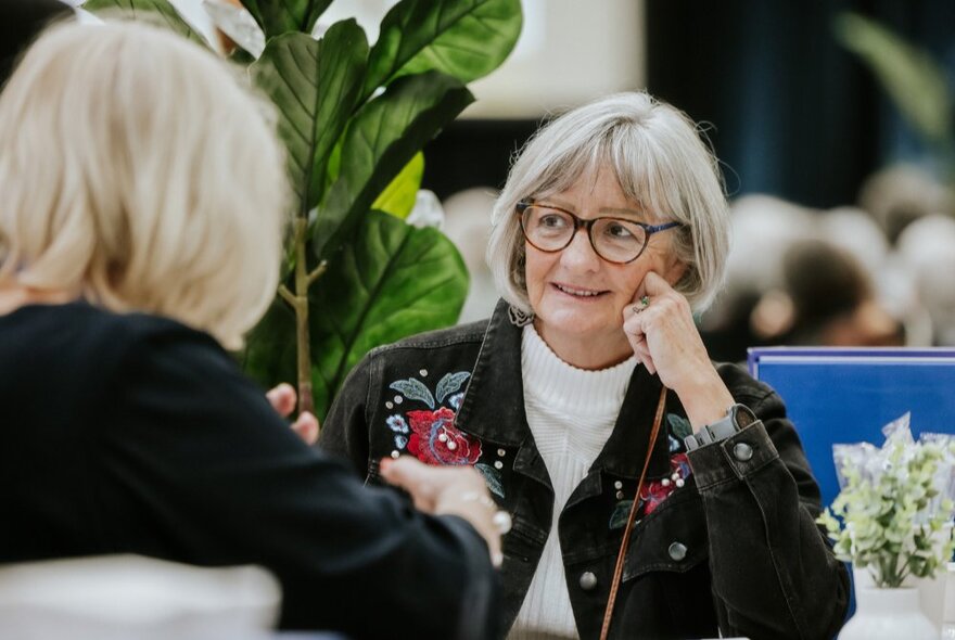 Two older people facing each other across a table talking, a green plant in the background.