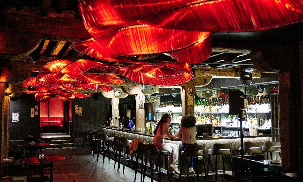 Dark and moody restaurant, dimly lit by a red ribbon light with two patrons sitting on stools at the bar.