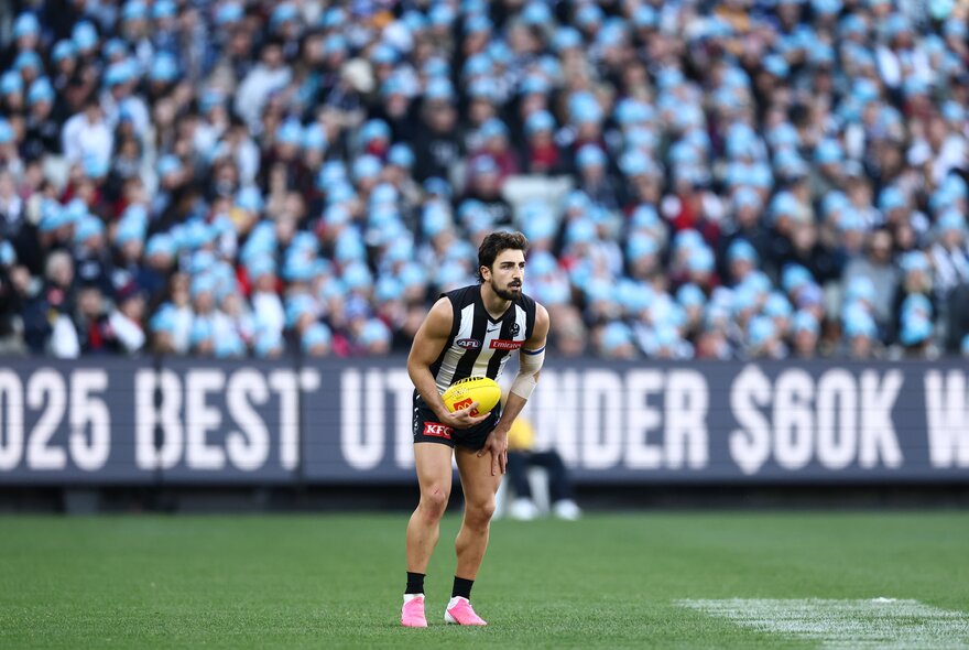 A Collingwood AFL player about to kick a yellow football he holds in his hands, with blurred fans in the stadium in the background.