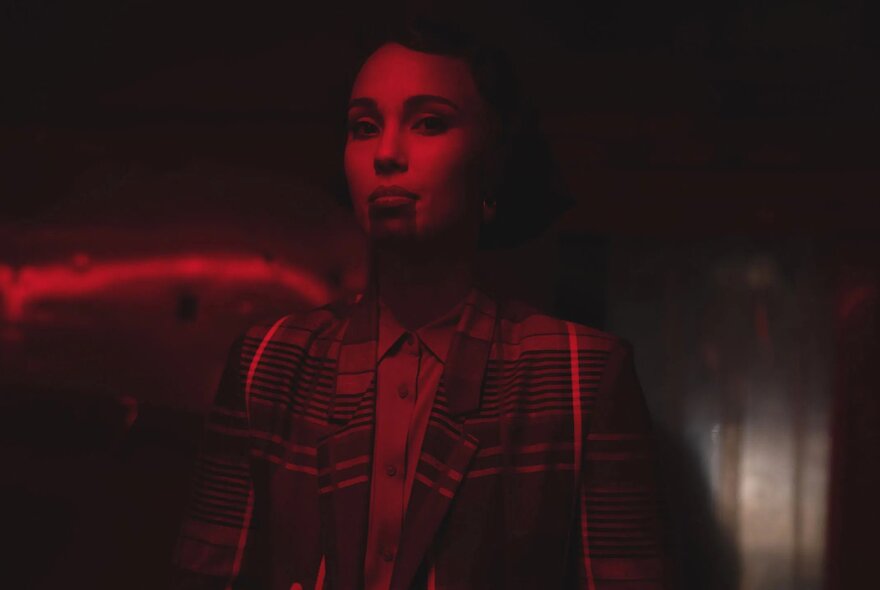 A still from the film BLKNWS: Terms & Conditions, depicts a person standing in a dimly lit room, a red wash over the image.