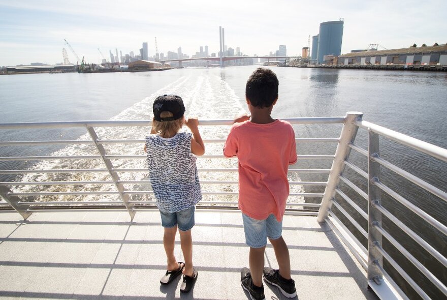 Two primary school aged children standing at the back of the large ferry looking out across the Yarra River, with the city skyline in the background.