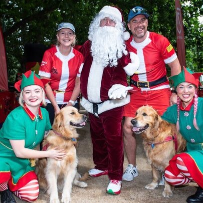 Two runners in Santa red t-shirts, posing for a photograph with Santa and two Elves and two dogs, outside in a park.