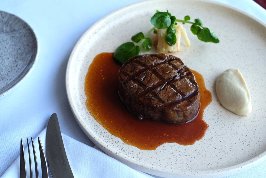 Restaurant-plated meal of steak and accompaniments on white napery with cutlery.