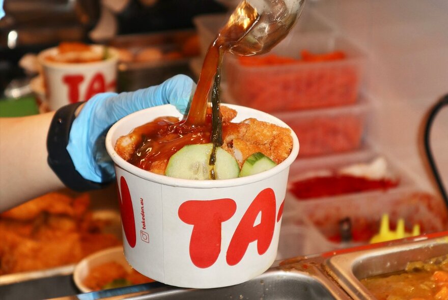 A paper cup being filled with a Japanese-Thai fusion takeaway dish.