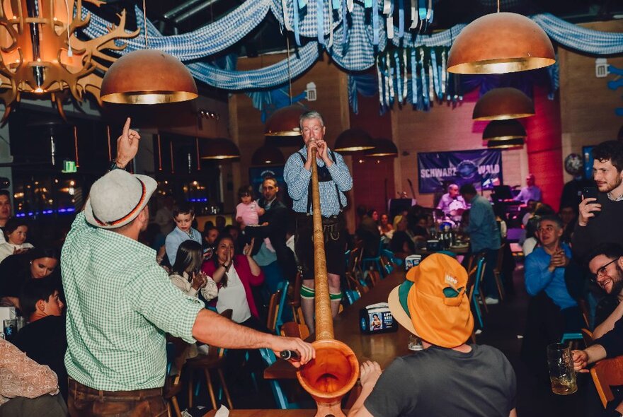 A man plays a very long horn in front of a room of people in a bar. 