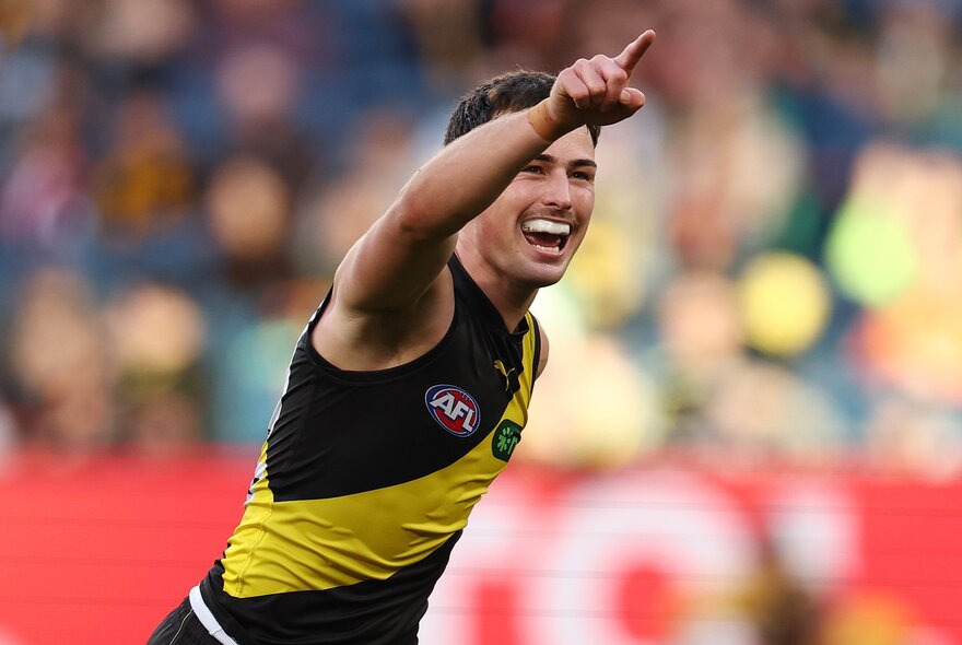 A Richmond AFL football player on the field during a match with his arm raised in victory, a blurred crowd of stadium fans in the background.