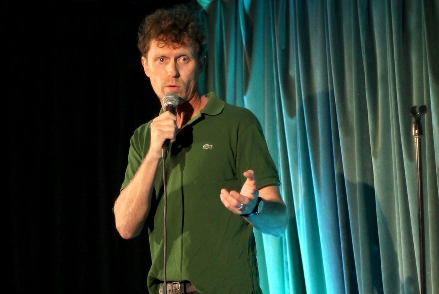 Comedian David Quirk on stage holding a microphone in his hand and performing stand up comedy.
