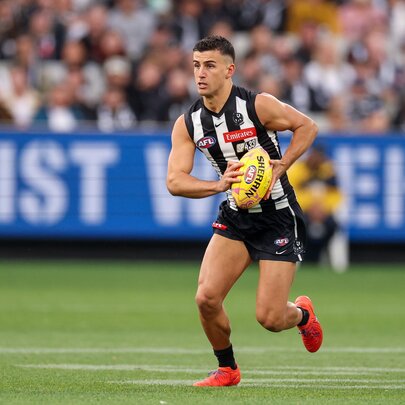 A Collingwood AFL football player running with the football during a match, with a blurred stadium crowd in the background.