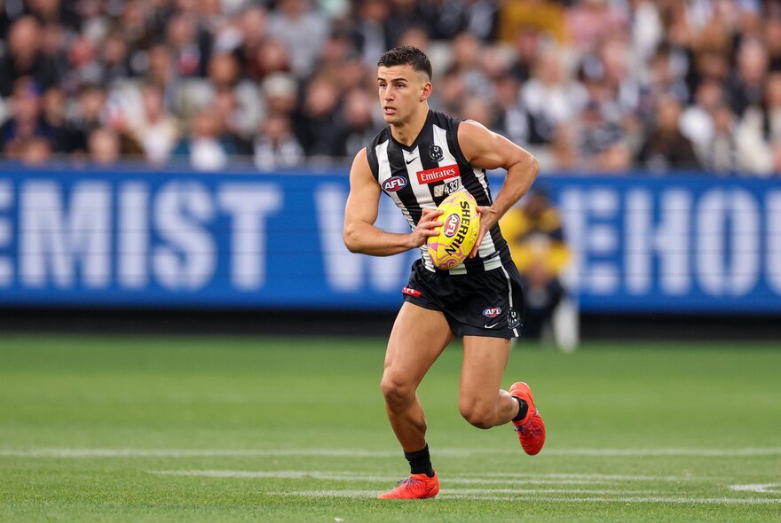 A Collingwood AFL football player running with the football during a match, with a blurred stadium crowd in the background.