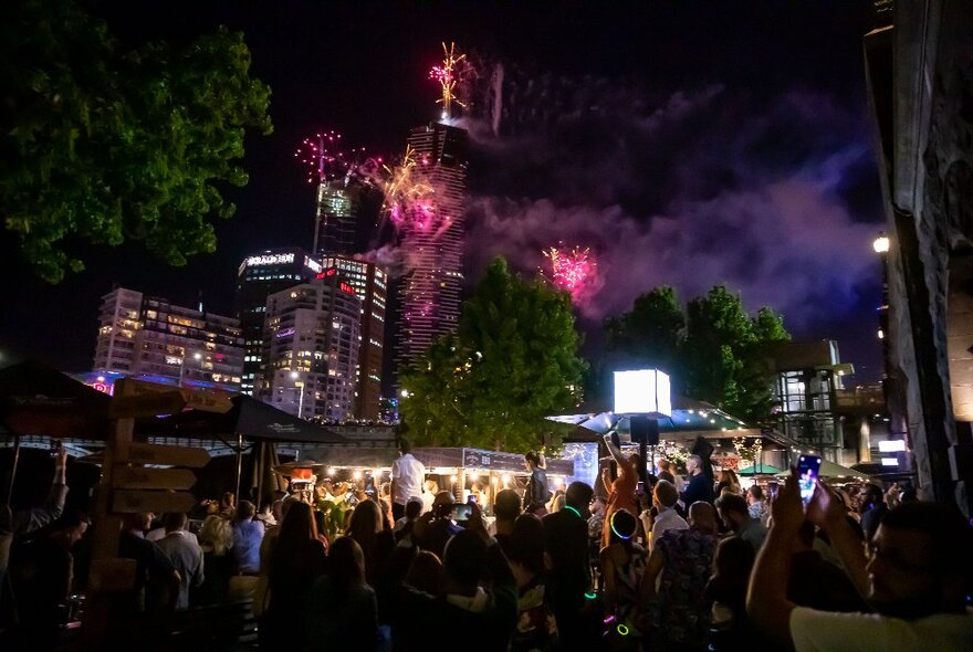 Crowded beer garden at night with smoke and pink fireworks in the sky.