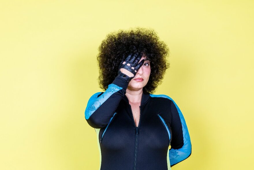 Person with an afro hairstyle stands in front of a pale lemon-yellow background and they are covering one eye with a leather-gloved hand.