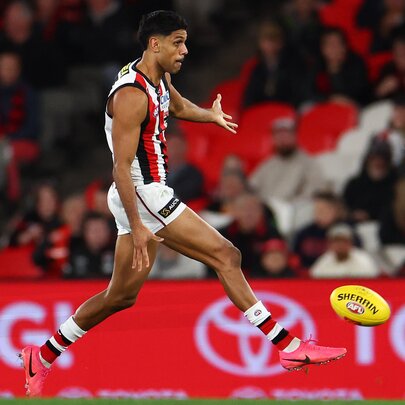 A St Kilda AFL player running while about to kick a yellow football he has just dropped in front of his leg, blurred fans in the stadium in the background.