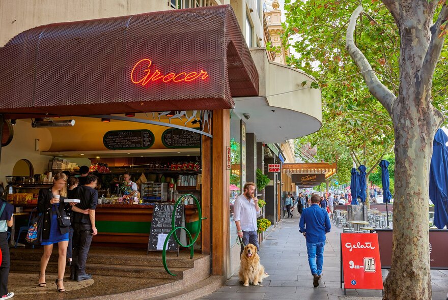 Outside a corner grocer and gelato shop. There is a man eating an ice cream and walking his dog. 