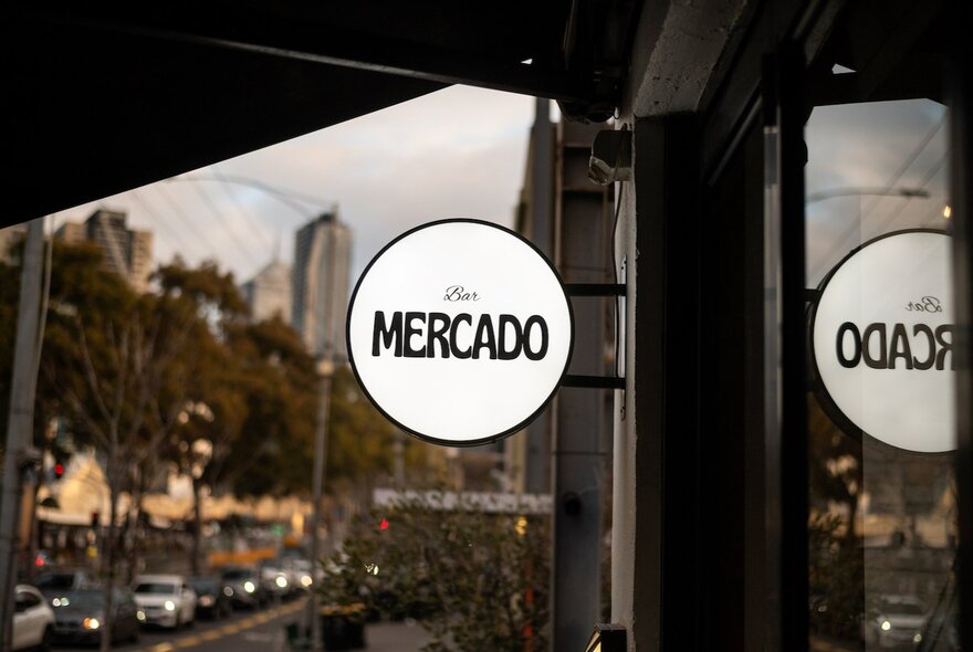 The exterior of Bar Mercado with a small illuminated round sign and the city's reflection in the front window.
