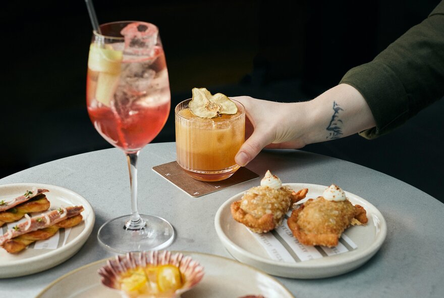 An hand with a tattoo on the wrist placing a cocktail down on a coaster on table with three small plates of snacks and a spritz. 