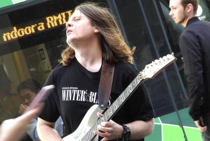 A busker performing in the street playing an electric guitar with his head of long hair tilted backwards and face expressing pain.