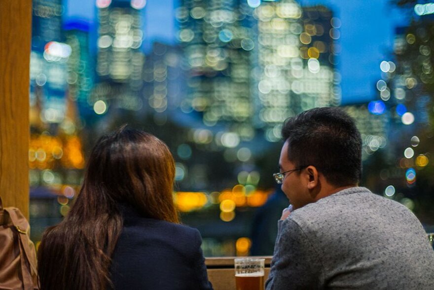 A couple enjoying a drink at a bar, with blurred city buildings at twilight in the background.