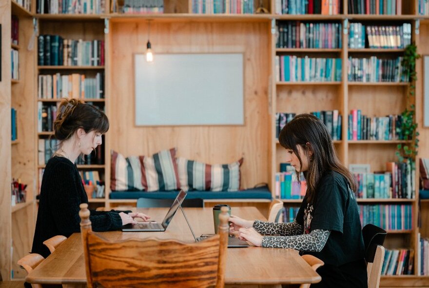 Two people sitting at a large table opposite each other and typing on their laptops, a wall of bookshelves behind them.