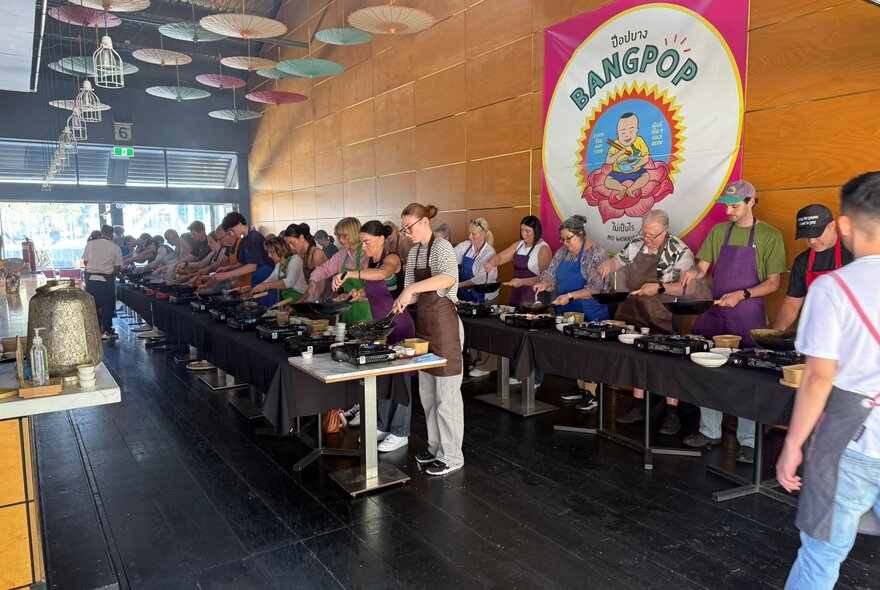 People lined up at workstations with Asian cooking equipment in a restaurant with ceiling hung with paper umbrellas.