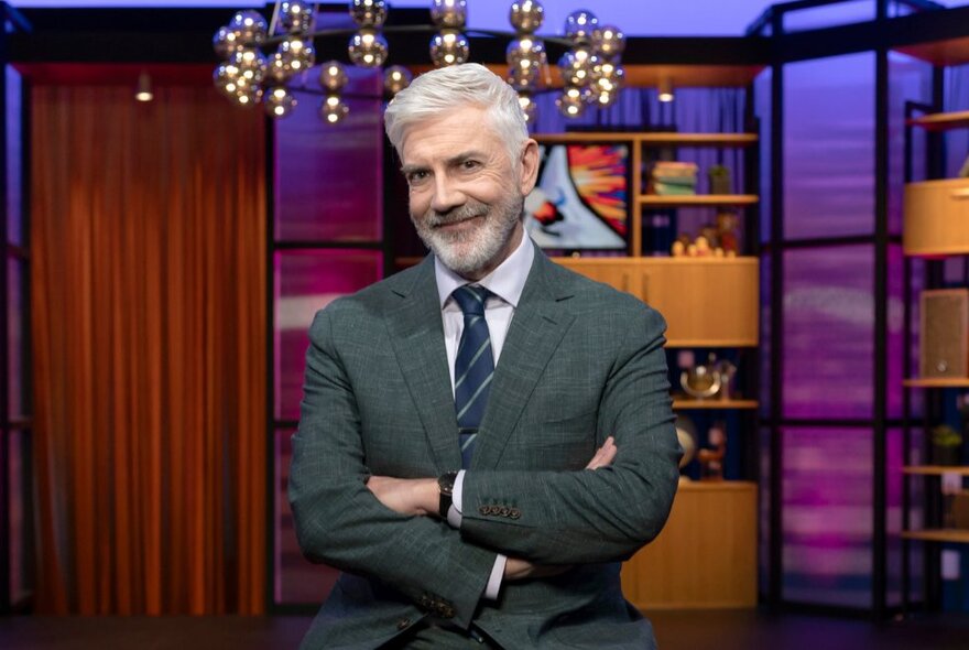 Television presenter Shaun Micallef wearing a suit, with his arms crossed in front of his chest, standing on the set of his TV chat show.