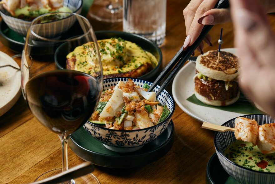 A table with a variety of dishes and a hand holding chopsticks, a glass of red wine in the foreground. 