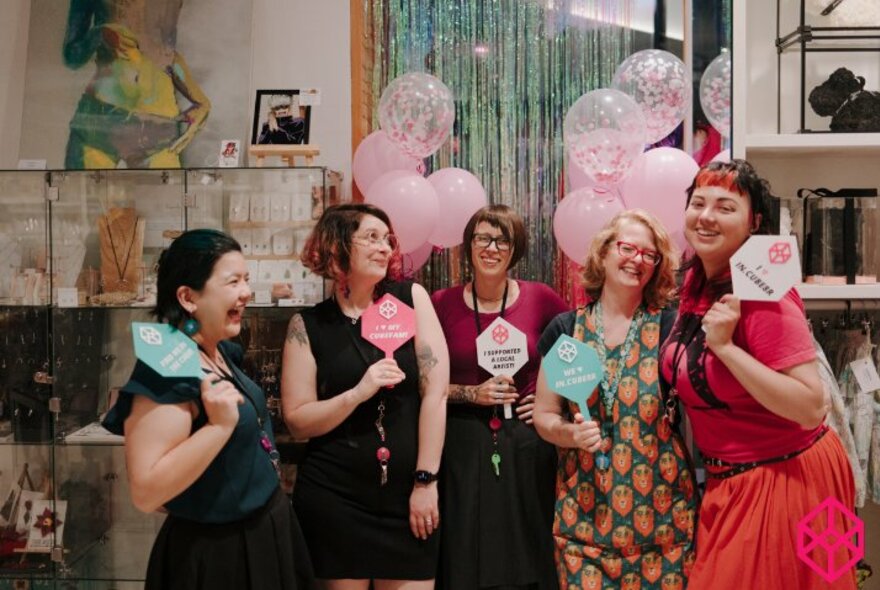 Group of five smiling people standing in a retail shop space, pink balloons behind them, all of them holding up small paper signs in their hands.