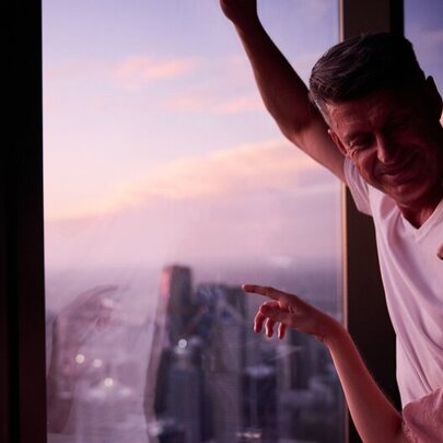 A couple standing at the window at Melbourne Skydeck and pointing out towards the Melbourne skyline at dusk.