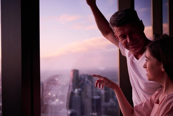 A couple standing at the window at Melbourne Skydeck and pointing out towards the Melbourne skyline at dusk.
