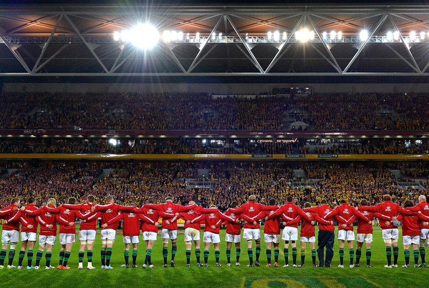 The Lions rugby team in red tops and white shorts, standing in a line with linked arms on a green sporting field in front of a stadium filled with fans.
