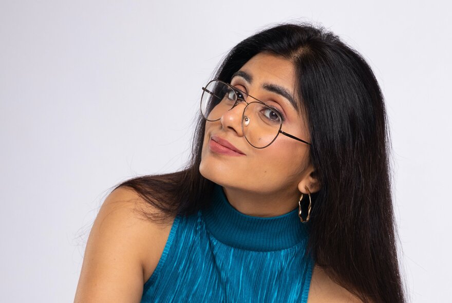 Indian comedian Prashasti Singh. She has long dark hair, is wearing round-rimmed glasses and a blue, textured, high-neck top. She is looking slightly upward and to the side with a smile. 