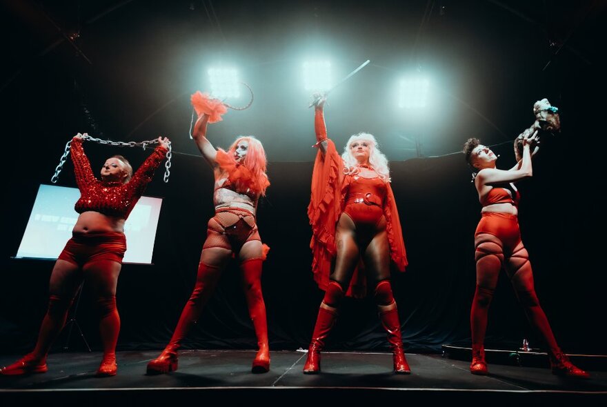 Four performers on a black stage, in red burlesque-circus style costumes and knee high red boots, with their arms raised in the air above them, spotlights behind them.