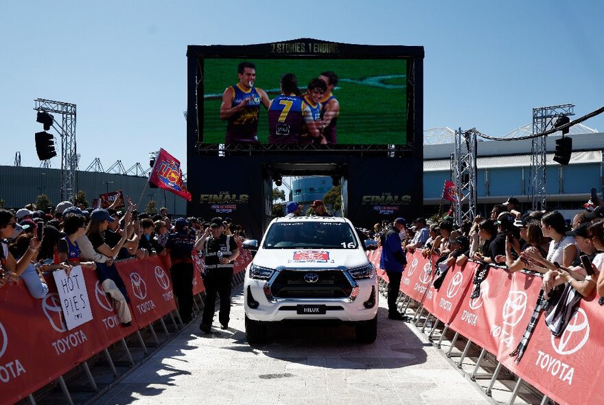 A Toyota Hilux makes its way through a crowd in the AFL Grand Final Parade with a giant TV screen behind. 