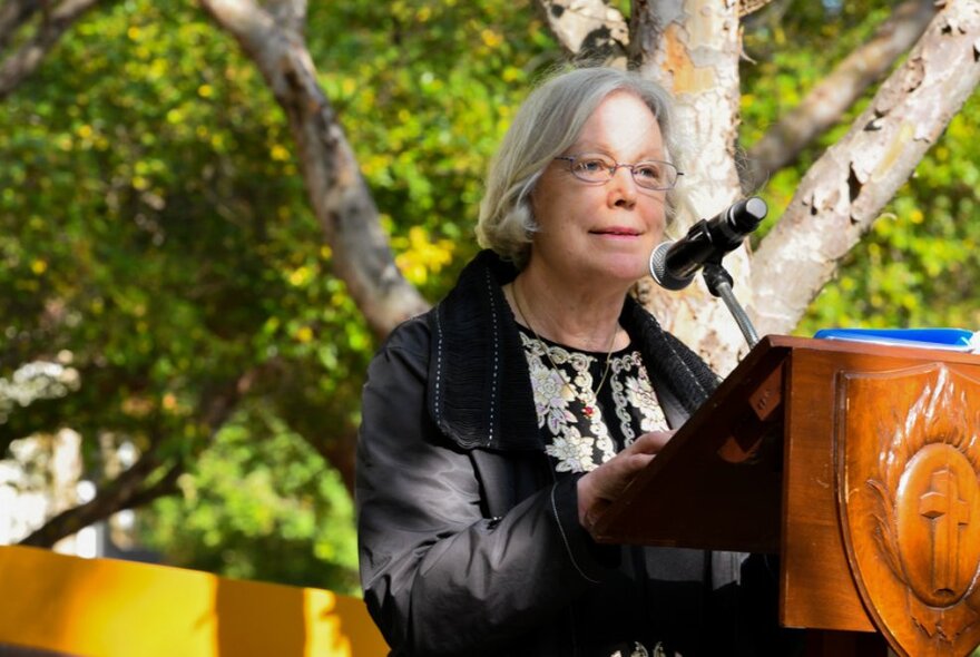 Author Judy Balcombe, a middle-aged woman with a grey bob haircut and glasses, standing at a lectern in an outdoor environment.