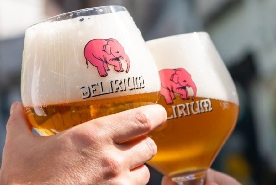 Hands holding two Belgian beers in large, wide beer glasses each with a pink elephant printed on it.