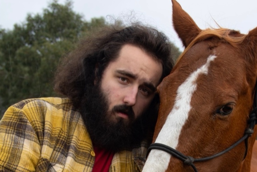 Man with beard and dark hair resting his head on the head of a horse, wearing a yellow plaid shirt.