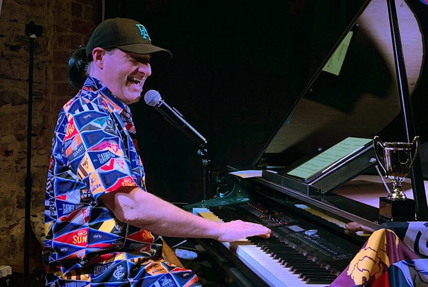 A man in a colourful shirt and baseball cap playing a black grand piano and singing into a microphone.