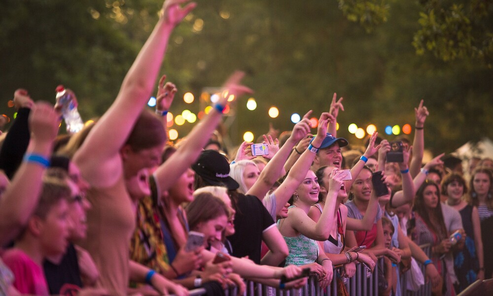 Packed crowd of people cheering, holding their hands up, and watching an event outside. 